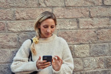 Çekici beyaz bir kadın cep telefonu kullanıyor. Gün boyunca dışarıda tuğla duvarın önünde dikiliyor.