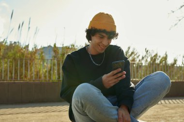 Güler yüzlü yakışıklı adam gün boyunca dışarıda bacak bacak üstüne atmış otururken cep telefonu kullanıyor.