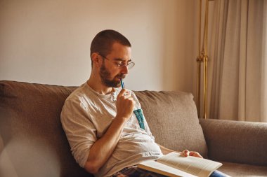 Düşünceli orta yaşlı bir adam oturuyor ve elinde bir kalemle kanepeye yaslanıyor.