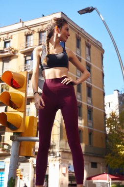 Arkasında büyük bir bina olan sarı bir trafik lambasının yanındaki duvarda duran koyu renk saçlı genç kadın.