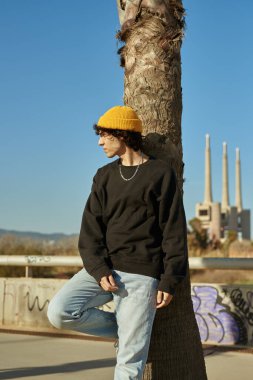 Yakışıklı, beyaz bir adam, gün boyunca ağaçlara yaslanırken dikkatle başka tarafa bakıyor.