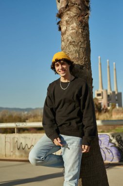 Güleryüzlü beyaz bir adam, gün boyunca ağaçlara yaslanırken kameraya bakıyor.