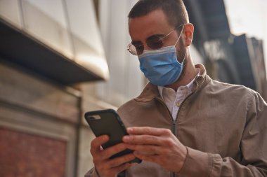 Dışarıda dikilirken maske takıp mesajlaşan dalgın bir adam.