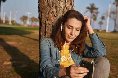 Genç bir kadın yaşlı bir ağacın yanında oturuyordu. Kulağında hoparlörler olan cep telefonuna bakıyordu.