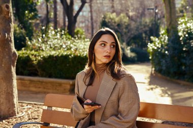 Kafası karışmış bir kadın bir elinde telefonuyla park yerinde oturuyor. Uzağa bakıyor.