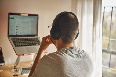 Dizüstü bilgisayar ve kulaklık kullanan nişanlı bir adam tek eliyle çenesini tutuyor.