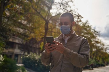 Büyülenmiş bir adam dışarıda duruyor ve iki eliyle mesaj yazıyor ve dikkatli bir şekilde maske takıyor.