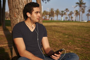 Çekici, koyu renk saçlı genç adam, telefonuna bağlı kulaklıklarını dinlerken uzaktan bakar.