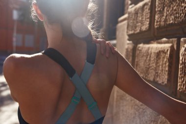 Kolunu omzuna dayamış, kaslarını esnetirken duvara destek veren bir kadının arka görüntüsü.