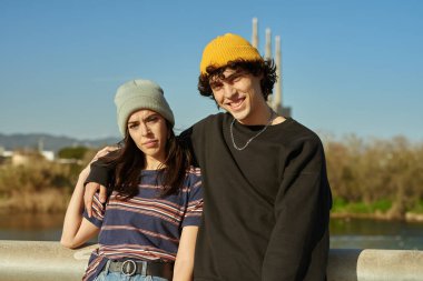 Yakışıklı, beyaz bir adam gün içinde köprüde koluyla sevgilisine sarılırken kameraya gülümsüyor.