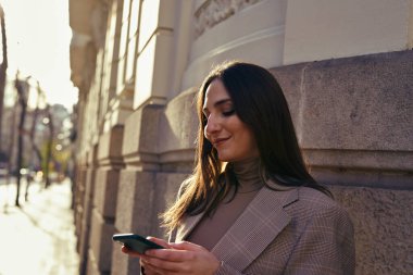 Bir binanın yanında duran sakin bayan gülümserken mesaj atıyor.