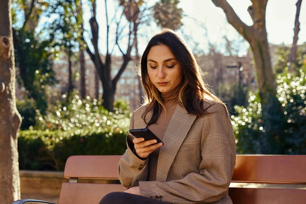 Intrigued lady sitting on a park bench with a straight posture while occupied with texting