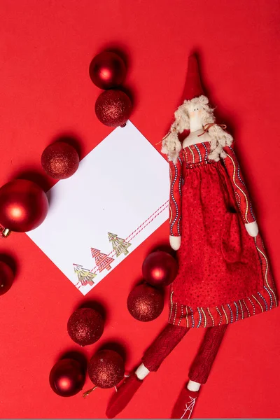 Blank Christmas card with rag doll on red table with Christmas baubles scattered all around in shades of red