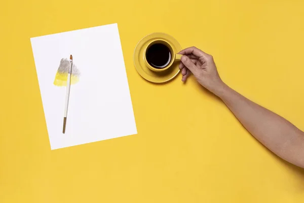 Hand holding yellow mug with hot coffee next to a white sheet for watercolor with gray and yellow color in gradient and brush