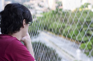 Siyah saçlı adam ve kırmızı tişört evcil hayvanlar için güvenlik ağı olan binanın balkonunda dalgın dalgın duruyor.