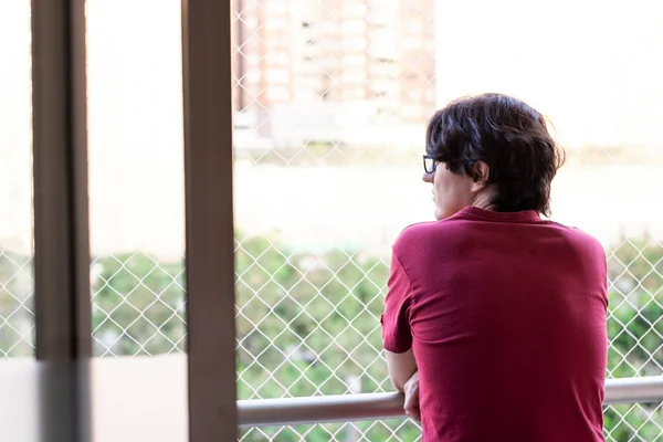 Kırmızı tişörtlü, siyah saçlı adam bir apartmanın balkonundan dışarı bakarken dalgın dalgın bakıyor.