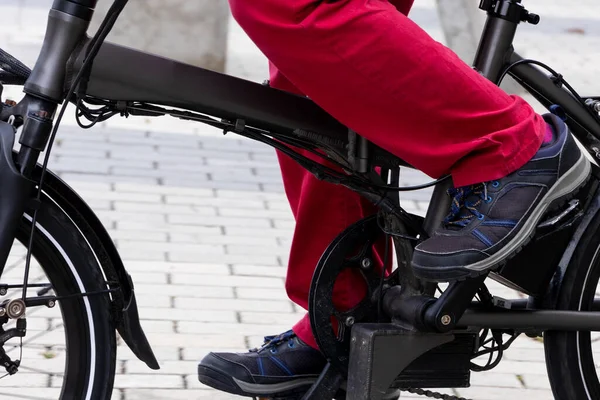 Kırmızı pantolonlu ve mavi pedallı siyah ayakkabılı birinin bacakları.