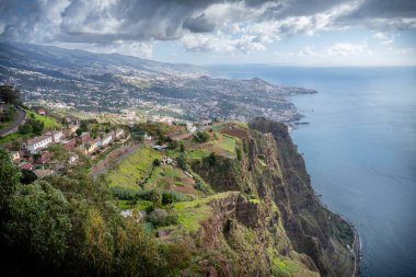 Madeira Adası 'ndaki Cabo Girao uçurumundan panoramik manzara Atlantik Okyanusu' nu, dağ evlerini ve dağınık bulutlu mavi gökyüzünün altındaki dramatik kıyı manzarasını gösteriyor..