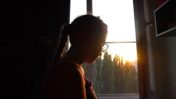 A teenage girl with glasses stands by the window. The light of the setting sun. Dark room. Green trees outside the window. Sad look. Close-up.