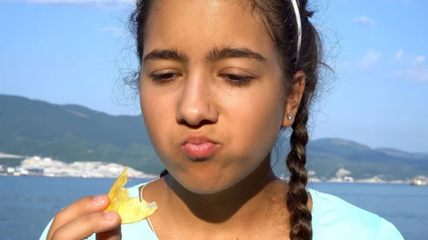 Teenage girl eating chips. Dark skin. Against the background of the seaport and mountains. The light of the sun at sunset. Blue sky. Close-up.