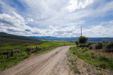 Çimenli bir arazinin ortasında sarı kapısı olan toprak bir yol. Gökyüzü bulutlu ve güneş parlıyor