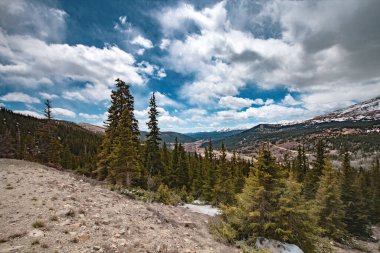 Açık mavi gökyüzü ve birkaç bulutu olan bir dağ sırası. Ağaçlar yeşil ve gökyüzü mavi.