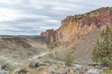 İçinden nehir akan kayalık bir dağ sırası. Gökyüzü bulutlu ve manzara çorak. Smith Rock State Park, Oregon, ABD.