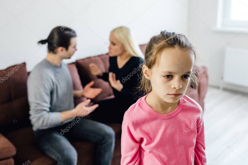 Frustrated little girl feeling depressed while angry parents fighting at home. Worried upset small daughter hurt by fathers and mothers break up or divorce, children and family conflict concept.