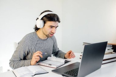 Erkek öğrenci kulaklık takıyor, uzaktan eğitim için laptop kullanıyor, konferans videosundan zevk alıyor, webinar izliyor, ses dersleri dinliyor, notlar yazıyor, öğretmenle çalışıyor, ders alıyor.