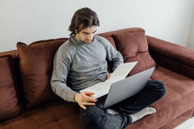 Evde dizüstü bilgisayar kullanan yakışıklı bir genç. Öğrenci adam kitabı okuyor. Çevrimiçi alışveriş, ev işi, serbest çalışma, çevrimiçi öğrenme, öğrenim kavramı. Uzaklık eğitimi