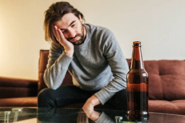 Güçlü baş ağrısı ya da migrenden muzdarip genç bir adam kanepeye oturmuş elinde şişeyle, milenyum adamı sarhoş ve ağrılı hissediyor, akşamdan kalma konseptinin ertesi sabahı
