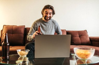 Çekici, neşeli, esmer adamın portresi evde dizüstü bilgisayar kullanarak divanda oturuyor. Film izlemek ya da online futbol maçı, işten sonra dinlenmek. yavaş çekim