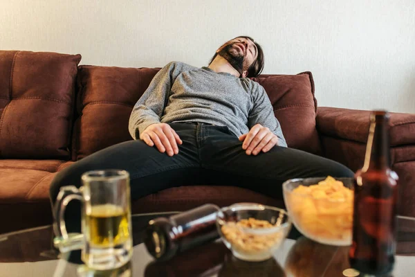喝醉了的年轻人在家里聚会后睡在沙发上 有食物和饮料的桌子是可见的 — 图库照片