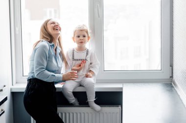 Küçük kızıyla birlikte, mutfaktaki pencerenin yanında oynayan mutlu beyaz Amerikalı anne. Ebeveynlik ve annelik Joy Concept. Anne ve kız evde seviniyor.