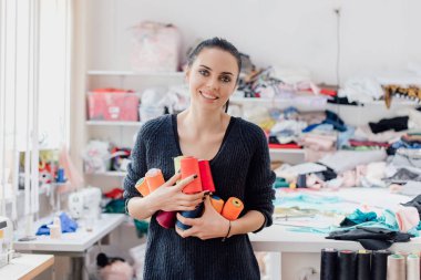  Şirin beyaz kadın terzi ya da terzi atölyenin arka planında renkli iplik makaraları tutuyor..