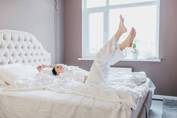 очаровательная, сексуальная, красивая, успешная, стильная брюнетка в пижаме, лежащая на спине с поднятыми ногами, наслаждающаяся временем дома в одиночестве, имеющая хорошее настроение по утрам