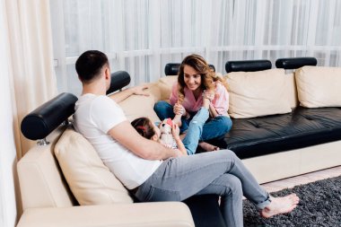 Mutlu genç anne ve baba ve küçük kızı kanepede oturuyor, küçük çocuk ve ailesi evde boş vakitlerin tadını çıkarıyor..