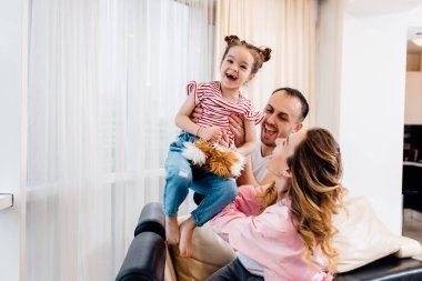 Mutlu küçük kız pencerenin yanındaki kanepede ailesiyle eğleniyor. Küçük çocuk ve ailesi evde oyun oynamaktan zevk alıyorlar..