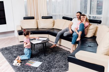 Küçük bir çocuk oturma odasında yere uzanmış renkli kalemlerle resim çiziyor, ebeveynler kanepede dinleniyor birlikte haftasonunun keyfini çıkarıyor, küçük çocuklar evde resim çiziyor, aile zamanı konsepti.