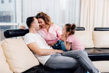 Çocuk ve ebeveynler arasındaki ilişki. Baba ve anne, kızları onlara yalvarırken gülümsüyor. Rahat kanepeye otur. Mutlu aile karantinada kalır..