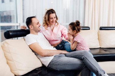 Çocuk ve ebeveynler arasındaki ilişki. Baba ve anne, kızları onlara yalvarırken gülümsüyor. Rahat kanepeye otur. Mutlu aile karantinada kalır..