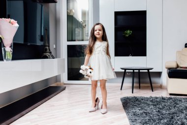 Beyaz elbiseli şirin küçük moda düşkünü. Mutlu kız çocuğu, annesinin ayakkabılarını ve kıyafetlerini oturma odasında dener. Gülümseyen komik küçük kız evde yürüyor annesinin topuklu ayakkabılarını deniyor.