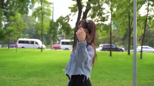 Vonzó és mosolygós tinédzser fejhallgatóval a fején. A parkban van., — Stock videók