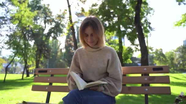 Krásná usměvavá dospívající dívka sedí na lavičce v parku a čtení knihy — Stock video
