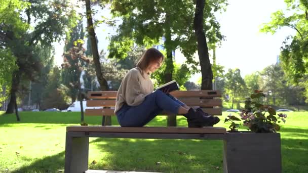 Hermosa adolescente sentada en un banco en el parque y leyendo libro. — Vídeo de stock