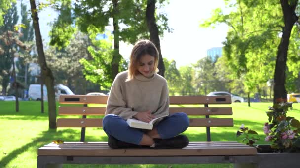Flicka sitter på en bänk i parken och läser bok. — Stockvideo