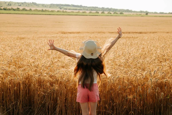 Vista trasera de niña pequeña con sombrero levantar las manos en tierra de trigo —  Fotos de Stock