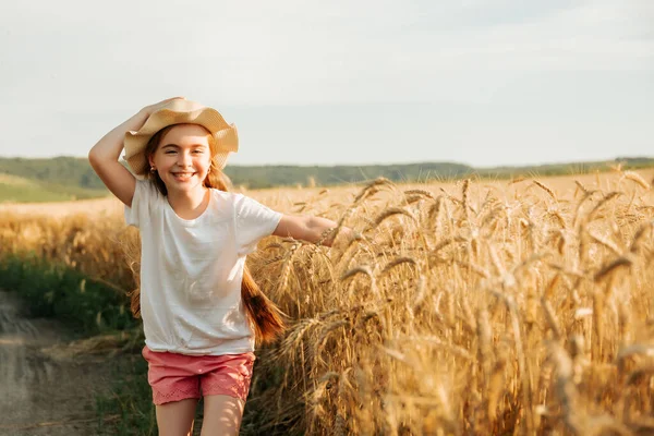 Adorable niña corre felizmente en el camino cerca del campo de trigo amarillo. —  Fotos de Stock