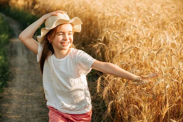Hermosa niña con sombrero en la cabeza y sonrisa atractiva corre por el camino del campo cerca del campo con trigo amarillo, —  Fotos de Stock
