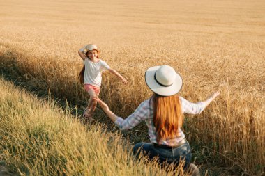 Kafasında şapkasıyla küçük mutlu bir kız annesine koşuyor buğday tarlasının yanında